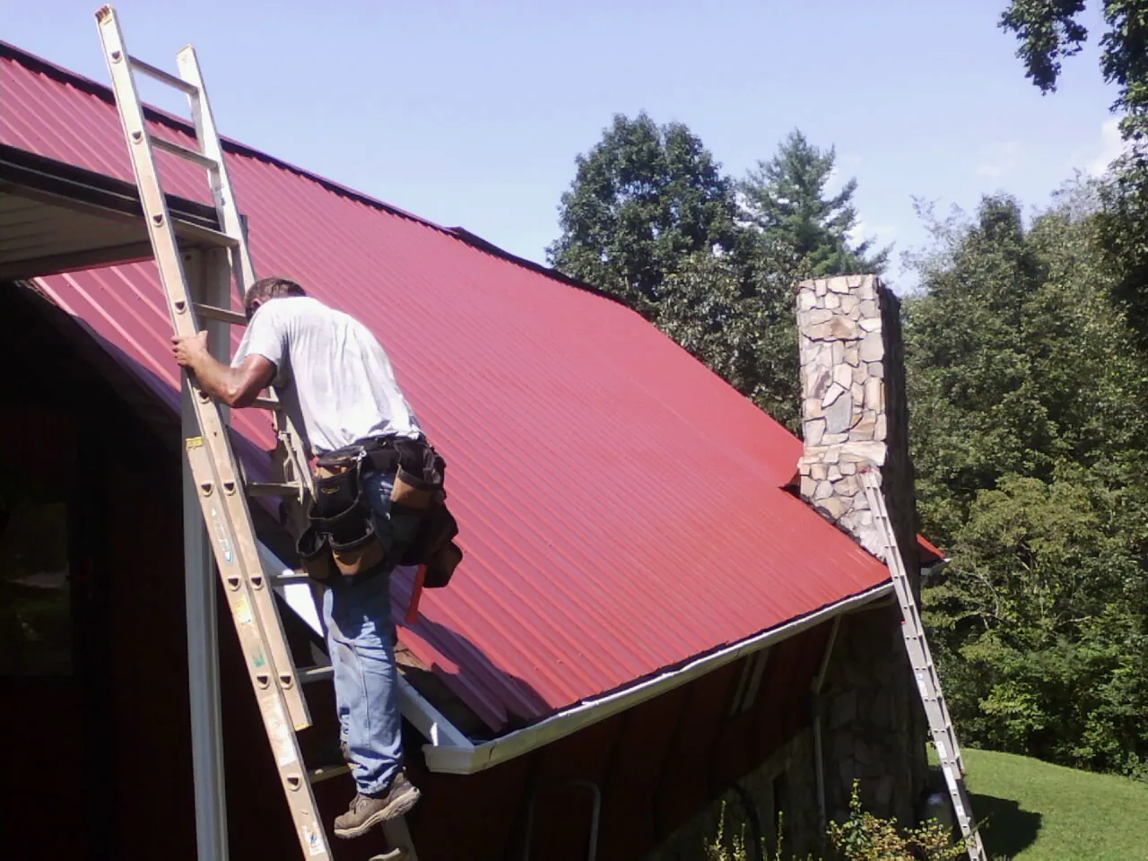 Roofer on a ladder beside a red metal roof and stone chimney — Soffit & Fascia Repair by Roofing Brothers in East Setauket